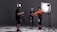 In a studio setting, three individuals are engaged in a photoshoot. One person is adjusting the attire of another, who is wearing a bright orange and black outfit, possibly for a motorsport or similar activity. The third person is checking a monitor or camera setup. The scene is illuminated by professional photography lighting equipment.