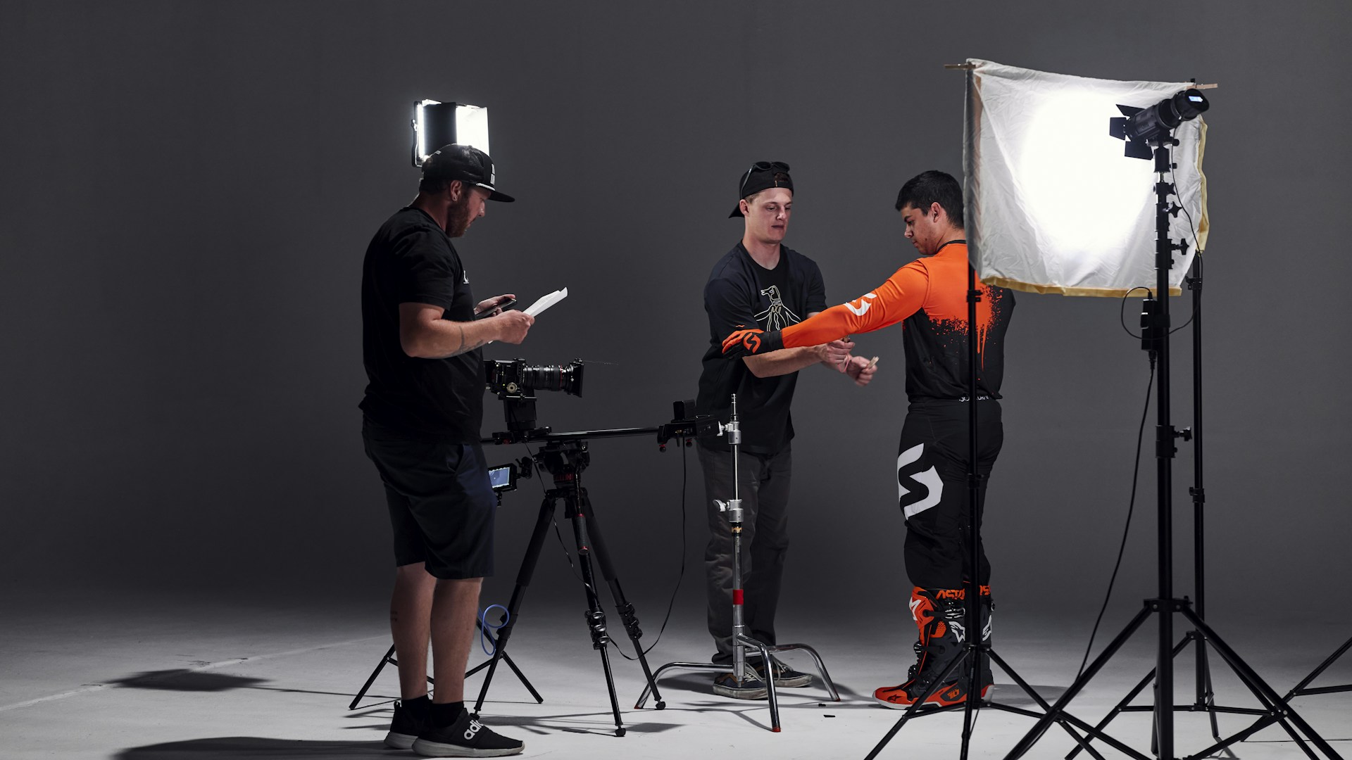 In a studio setting, three individuals are engaged in a photoshoot. One person is adjusting the attire of another, who is wearing a bright orange and black outfit, possibly for a motorsport or similar activity. The third person is checking a monitor or camera setup. The scene is illuminated by professional photography lighting equipment.