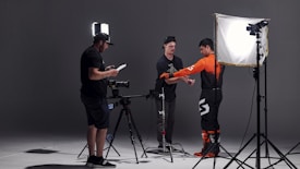 In a studio setting, three individuals are engaged in a photoshoot. One person is adjusting the attire of another, who is wearing a bright orange and black outfit, possibly for a motorsport or similar activity. The third person is checking a monitor or camera setup. The scene is illuminated by professional photography lighting equipment.