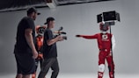 A group of individuals in a studio setting are engaged in what appears to be a filming or photoshoot session. Two people are wearing protective racing gear and helmets, while one person is holding a camera mounted on a stabilizer. Another individual is present, seemingly observing or directing the scene. The environment is well-lit with the use of studio lights, creating a focused and professional atmosphere.