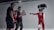 A group of individuals in a studio setting are engaged in what appears to be a filming or photoshoot session. Two people are wearing protective racing gear and helmets, while one person is holding a camera mounted on a stabilizer. Another individual is present, seemingly observing or directing the scene. The environment is well-lit with the use of studio lights, creating a focused and professional atmosphere.