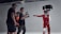 A group of individuals in a studio setting are engaged in what appears to be a filming or photoshoot session. Two people are wearing protective racing gear and helmets, while one person is holding a camera mounted on a stabilizer. Another individual is present, seemingly observing or directing the scene. The environment is well-lit with the use of studio lights, creating a focused and professional atmosphere.