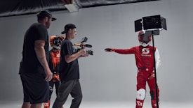 A group of individuals in a studio setting are engaged in what appears to be a filming or photoshoot session. Two people are wearing protective racing gear and helmets, while one person is holding a camera mounted on a stabilizer. Another individual is present, seemingly observing or directing the scene. The environment is well-lit with the use of studio lights, creating a focused and professional atmosphere.