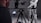 A person operates a professional camera setup on a tripod, with a monitor displaying an image. The camera is pointed at a person wearing a black and white racing suit, which includes logos and text. The background is a neutral, dark gray color.