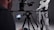 A person operates a professional camera setup on a tripod, with a monitor displaying an image. The camera is pointed at a person wearing a black and white racing suit, which includes logos and text. The background is a neutral, dark gray color.