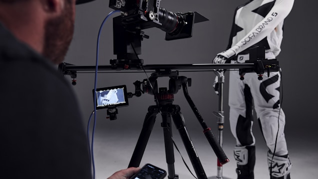 A person operates a professional camera setup on a tripod, with a monitor displaying an image. The camera is pointed at a person wearing a black and white racing suit, which includes logos and text. The background is a neutral, dark gray color.