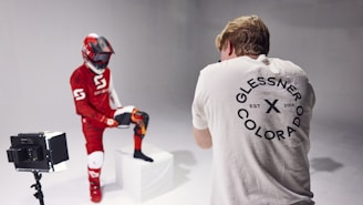 A person wearing a red and white motocross outfit with a helmet poses with one leg on a white block in a photography studio. Another person in a white shirt with a circular logo on the back stands in front of the camera, taking photographs. There is a light box to the left of the person in the outfit.