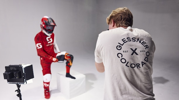 A person wearing a red and white motocross outfit with a helmet poses with one leg on a white block in a photography studio. Another person in a white shirt with a circular logo on the back stands in front of the camera, taking photographs. There is a light box to the left of the person in the outfit.