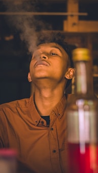 A young person wearing a brown shirt is leaning back slightly while exhaling smoke. The scene has a warm, indoor ambiance with hints of wood in the background. A blurred bottle is in the foreground.