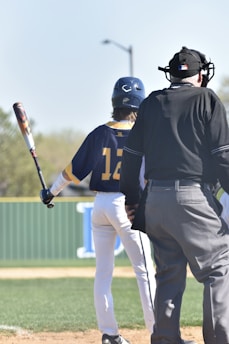 A baseball player wearing a helmet and uniform with the number 12 stands at bat, holding a baseball bat. An umpire is positioned nearby, dressed in a black uniform with protective gear. The setting is an outdoor baseball field with a green fence and natural surroundings.