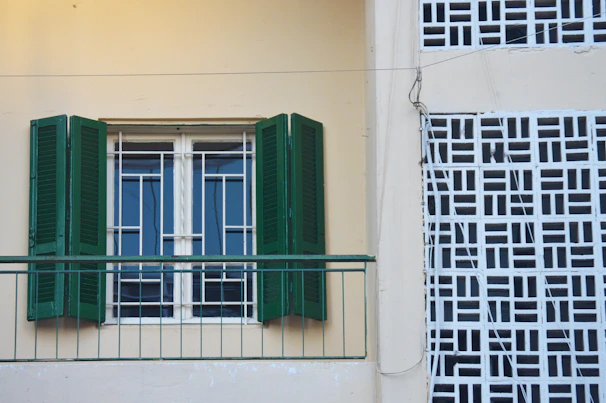 A building facade featuring a window with green shutters and a metal railing. The wall is light yellow, and adjacent to the window is a decorative lattice pattern in white with black gaps.