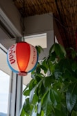 A paper lantern with red, white, and blue horizontal stripes and some Japanese writing hangs next to a green leafy plant by a window. The lantern emits a warm glow, and the plant provides a natural contrast with its vibrant green leaves. Above, there is a ceiling made of bamboo material, adding to the organic ambiance.