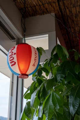 A paper lantern with red, white, and blue horizontal stripes and some Japanese writing hangs next to a green leafy plant by a window. The lantern emits a warm glow, and the plant provides a natural contrast with its vibrant green leaves. Above, there is a ceiling made of bamboo material, adding to the organic ambiance.