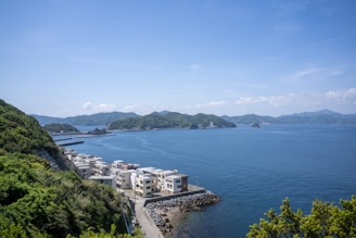 A coastal scene featuring a series of modern buildings near the edge of a calm sea. The buildings are situated along a narrow road that runs parallel to the shoreline, with a rocky embankment separating them from the water. Lush green hills envelop the left side of the image, while distant forested islands can be seen across the water under a clear, blue sky.