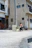 A person walks along a quiet urban street carrying a small dog. The area is surrounded by residential buildings, featuring a mix of architectural styles and mostly neutral colors. A bicycle is parked near a utility pole, and the street is mostly empty.