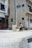 A person walks along a quiet urban street carrying a small dog. The area is surrounded by residential buildings, featuring a mix of architectural styles and mostly neutral colors. A bicycle is parked near a utility pole, and the street is mostly empty.