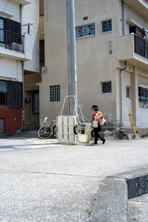 A person walks along a quiet urban street carrying a small dog. The area is surrounded by residential buildings, featuring a mix of architectural styles and mostly neutral colors. A bicycle is parked near a utility pole, and the street is mostly empty.