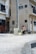 A person walks along a quiet urban street carrying a small dog. The area is surrounded by residential buildings, featuring a mix of architectural styles and mostly neutral colors. A bicycle is parked near a utility pole, and the street is mostly empty.