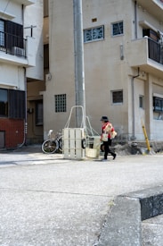 A person walks along a quiet urban street carrying a small dog. The area is surrounded by residential buildings, featuring a mix of architectural styles and mostly neutral colors. A bicycle is parked near a utility pole, and the street is mostly empty.