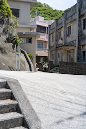 An urban alleyway flanked by concrete buildings, with visible stairs on the left and greenery on top of a rocky surface. In the distance, a person is walking while carrying bags. The atmosphere is serene and the setting appears quiet and somewhat deserted.