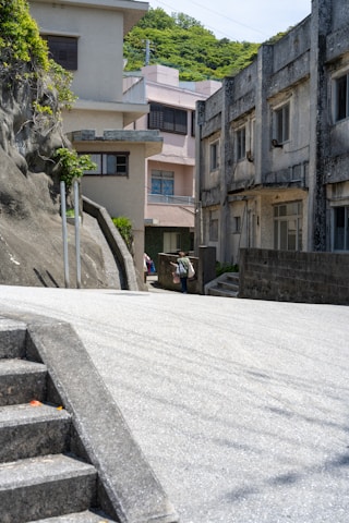 An urban alleyway flanked by concrete buildings, with visible stairs on the left and greenery on top of a rocky surface. In the distance, a person is walking while carrying bags. The atmosphere is serene and the setting appears quiet and somewhat deserted.