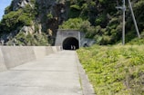 A paved pathway leads up to a tunnel entrance set within a rocky hillside. Lush green vegetation covers the surrounding landscape. Three people can be seen near the tunnel entrance. Utility poles with overhead wires run along the right side of the path.