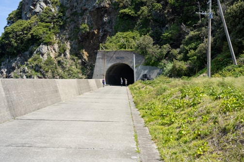A paved pathway leads up to a tunnel entrance set within a rocky hillside. Lush green vegetation covers the surrounding landscape. Three people can be seen near the tunnel entrance. Utility poles with overhead wires run along the right side of the path.