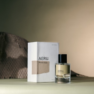 A perfume bottle and its accompanying packaging are arranged on a surface with a muted, textured background. The perfume bottle has a sleek, minimalist design with a black cap and is labeled 'AERU. Blossom Island'. The packaging box is simple, featuring similar branding and a fabric-like detail.