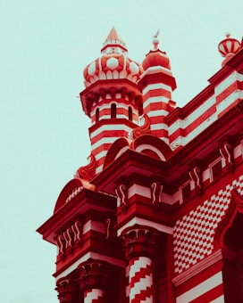 An architectural structure featuring intricate red and white striped patterns. The building has tall, ornate towers with domed tops and detailed carvings. The architectural style appears elaborate and grandiose, with a mix of cylindrical and rectangular elements.