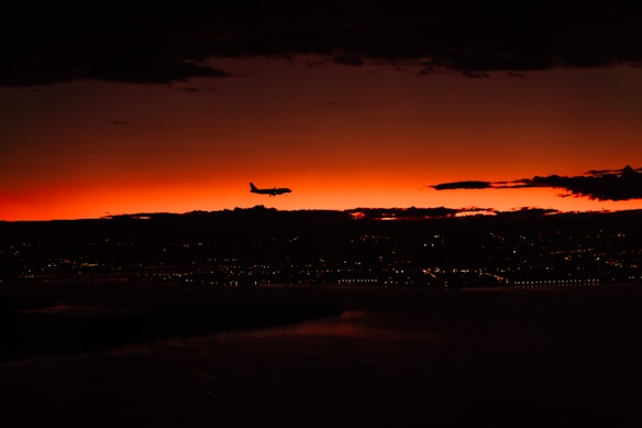 A silhouette of an airplane is flying against a vibrant sunset sky, which is a deep red and orange. Below the airplane, faint outlines of a cityscape can be seen, with scattered lights twinkling above a body of water, creating a tranquil evening scene.