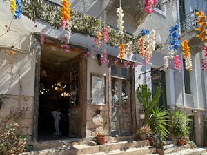 A charming storefront decorated with vibrant hanging flowers in shades of blue, pink, orange, and white. The entrance features an ornate, rustic door surrounded by lush potted plants including cacti and other greenery. A statue is visible inside, along with warm lighting fixtures and a sign indicating the space is open.