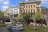 A canal scene with a blue tour boat and a smaller boat on the water, surrounded by historic buildings with a mix of architectural styles. The area is lively with people enjoying outdoor dining under red umbrellas. Trees add greenery to the scene.