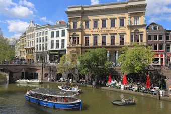 A canal scene with a blue tour boat and a smaller boat on the water, surrounded by historic buildings with a mix of architectural styles. The area is lively with people enjoying outdoor dining under red umbrellas. Trees add greenery to the scene.