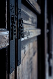 Close-up view of an aged wooden door featuring intricate metal hinges with a rustic texture. The wood displays visible grain and wear, while the metal hinges are adorned with a cut-out pattern, suggesting a blend of traditional craftsmanship.