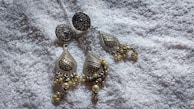 A pair of intricately designed metallic earrings with teardrop shapes, featuring detailed patterns and decorative elements like small pearls. They are placed on a soft, textured white fabric that provides a contrasting background.