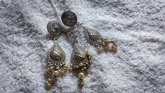 A pair of intricately designed metallic earrings with teardrop shapes, featuring detailed patterns and decorative elements like small pearls. They are placed on a soft, textured white fabric that provides a contrasting background.