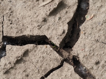 A close-up view of cracked and dry soil with large fissures. The soil appears parched and crumbly, with some small pebbles and a few tiny sprouts of green vegetation emerging from the cracks. A single dried leaf is visible on the top right corner of the image.