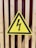A yellow triangular warning sign with a black border displays a black lightning bolt symbol, indicating electrical hazard. It is mounted on a wooden slatted surface with a red background visible through the slats.