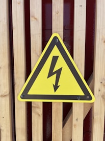 A yellow triangular warning sign with a black border displays a black lightning bolt symbol, indicating electrical hazard. It is mounted on a wooden slatted surface with a red background visible through the slats.
