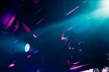 An industrial or stage setting is illuminated by dramatic lighting. Beams of blue and purple light create a futuristic and moody atmosphere against a backdrop of structural elements, possibly in a warehouse or theater.