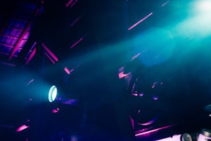 An industrial or stage setting is illuminated by dramatic lighting. Beams of blue and purple light create a futuristic and moody atmosphere against a backdrop of structural elements, possibly in a warehouse or theater.