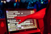 A person's hand interacts with a touchscreen device displaying a complex interface with multiple squares and labels. The scene is illuminated with red light, suggesting a vibrant and dynamic environment.
