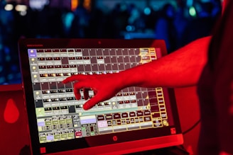 A person's hand interacts with a touchscreen device displaying a complex interface with multiple squares and labels. The scene is illuminated with red light, suggesting a vibrant and dynamic environment.