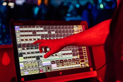 A person's hand interacts with a touchscreen device displaying a complex interface with multiple squares and labels. The scene is illuminated with red light, suggesting a vibrant and dynamic environment.