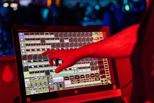 A person's hand interacts with a touchscreen device displaying a complex interface with multiple squares and labels. The scene is illuminated with red light, suggesting a vibrant and dynamic environment.