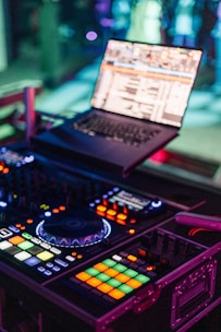 A DJ setup with a laptop displaying music software and a colorful, illuminated DJ controller. The image captures the vibrant atmosphere of a music venue, with glowing buttons and knobs on the controller.