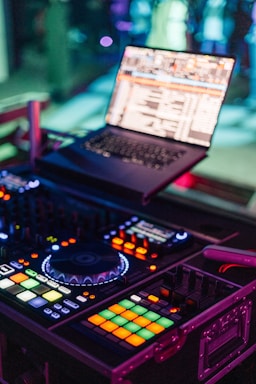 A DJ setup with a laptop displaying music software and a colorful, illuminated DJ controller. The image captures the vibrant atmosphere of a music venue, with glowing buttons and knobs on the controller.