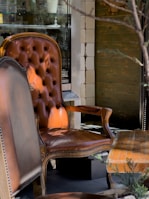 A vintage leather armchair with a tufted backrest placed in an outdoor setting. The armchair has a rich brown color with ornate wooden arms and legs. Next to it, there is a matching wooden table with an upholstered seat, and the scene is set against a rustic wooden wall with visible textures. The lighting casts soft shadows, adding depth to the overall composition.