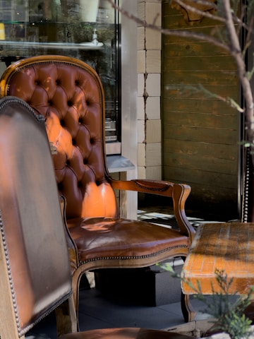 A vintage leather armchair with a tufted backrest placed in an outdoor setting. The armchair has a rich brown color with ornate wooden arms and legs. Next to it, there is a matching wooden table with an upholstered seat, and the scene is set against a rustic wooden wall with visible textures. The lighting casts soft shadows, adding depth to the overall composition.