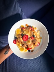 A bowl of cereal topped with slices of banana and strawberries, blueberries, almonds, and a sprinkle of what appears to be cinnamon or another spice. The bowl is white and held by a person with a visible arm tattoo against a blue fabric background.